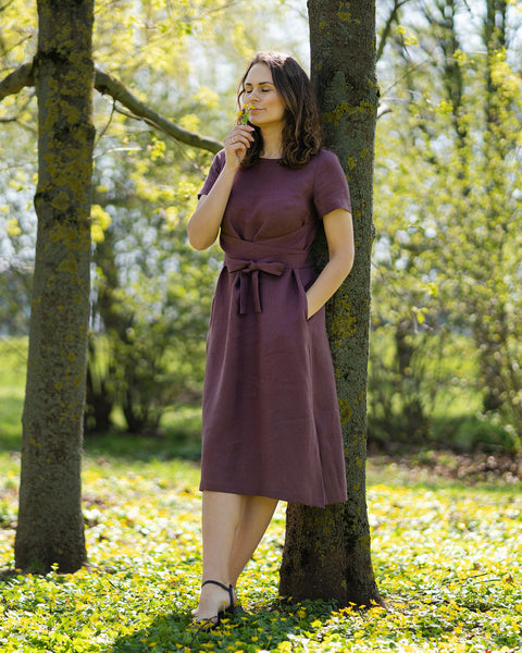 A woman with brown hair, wearing a short-sleeved purple dress, stands outdoors on the grass among small yellow flowers.
