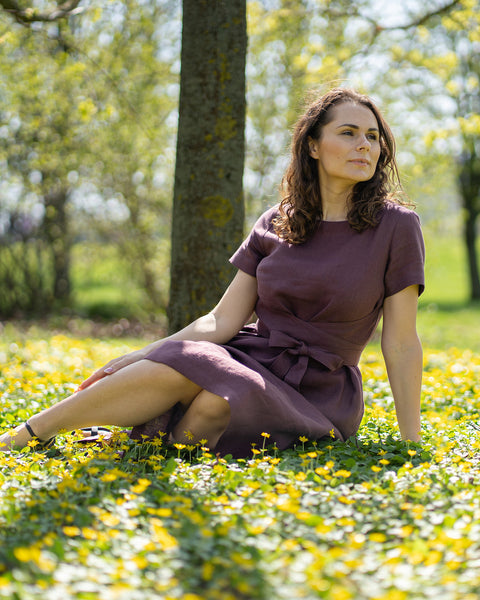 A woman with brown hair, wearing a short-sleeved purple dress, sits outdoors on the grass among small yellow flowers.
