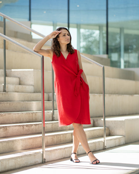 A woman with brown hair, wearing a bright red, sleeveless wrap dress, poses on indoor concrete stairs with metal railings.
