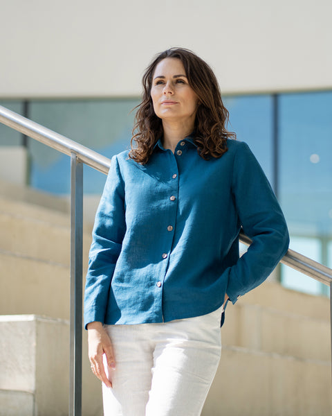 A woman with brown hair, wearing a cobalt blue button-front shirt and wide-leg white trousers, stands outdoors against a modern concrete structure.