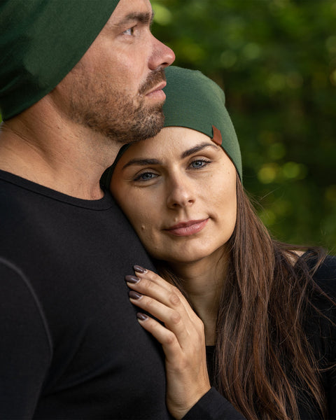 A picture featuring a couple wearing the unisex dark green merino beanie. It has a small logo on the side at the front. The woman is resting her head and palm on the man&#39;s chest. He is looking to the side.