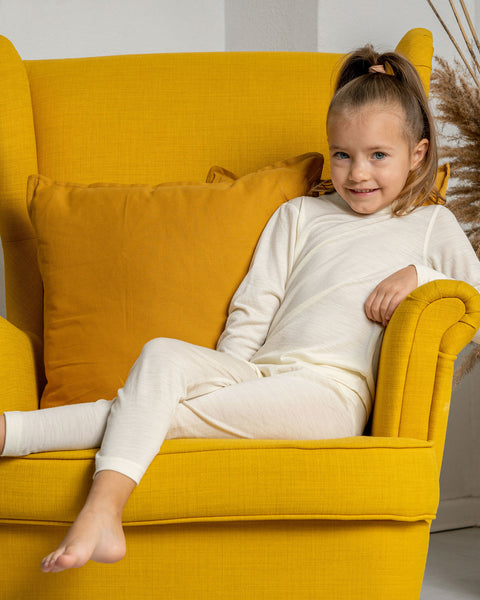 A girl sitting in an armchair in yellow color, wearing the menique kids&#39; pants from merino fabric in natural color. 