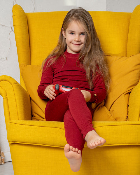 A girl, sitting in a yellow armchair, holding a game controller wearing the menique Kids&#39; 160 Merino Pants in Royal Cherry color. 