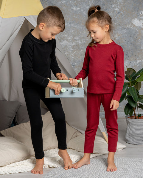 Two young children in thermal base layer sets — the boy in black and the girl in red — playing with a wooden toy together in a cozy indoor setting with a play tent, cushions, and potted plants.