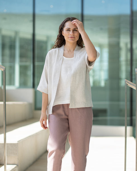 A woman with brown hair, wearing a menique natural kimono-style jacket over mauve trousers, stands on indoor concrete stairs.
