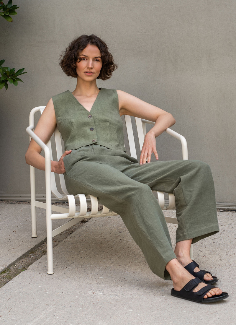 Woman wearing a sleeveless olive green linen vest and matching wide-leg linen trousers, sitting relaxed on a white metal chair against a neutral wall. The outfit features a tailored button-front vest and breathable natural fabric, styled with black sandals for a minimalist, modern summer look.