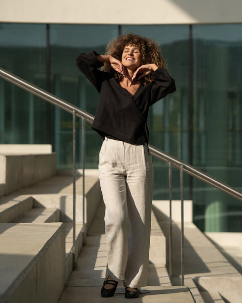 A smiling woman with voluminous curly brown hair stands on concrete steps, her hands framing her face. She is wearing a pure black long-sleeved wrap top and menique linen, wide-legged pants, paired with black ballet-style shoes with straps. The background features a modern building with large, reflective windows and a metal railing along the steps, suggesting an urban or architectural setting in bright sunlight.