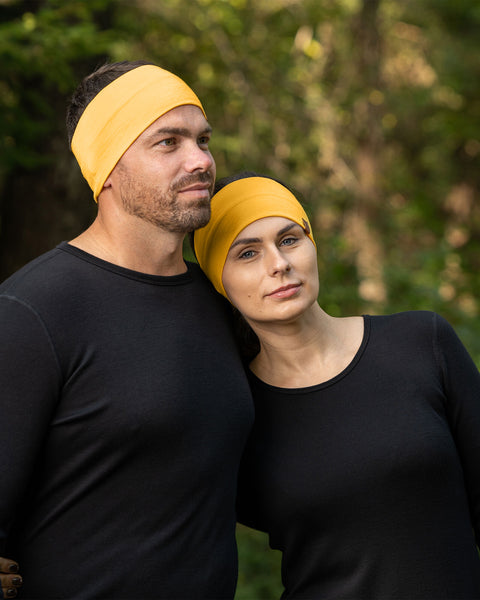 A man and a woman are standing close together outdoors, both wearing menique spicy yellow headbands and black long-sleeved shirts. The man has short brown hair and a beard, and he is looking slightly upwards and to the left. 