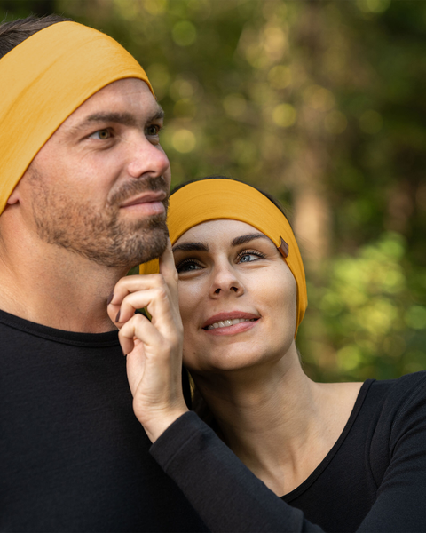 A man and a woman are closely positioned outdoors, both wearing spicy yellow menique merino wool headbands and black long-sleeved shirts. The man, on the left, has short brown hair and a beard and is looking upwards and to his left. The woman, on the right, has light brown hair and is looking upwards with a smile, her hand gently touching the man's face. The background is softly blurred with green and brown tones, suggesting a natural, possibly wooded, environment.