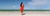 A woman with long dark hair stands on a sandy beach, facing away from the camera. She wears a long-sleeved orange dress and is barefoot. The ocean is in the background with light waves, and the sky is blue with a few clouds.