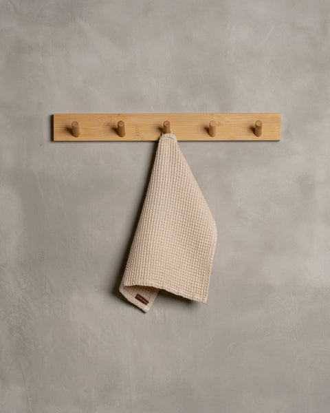 A gray wall with a wooden hanger. In the middle of it hangs a waffle face washcloth in sand brown color.