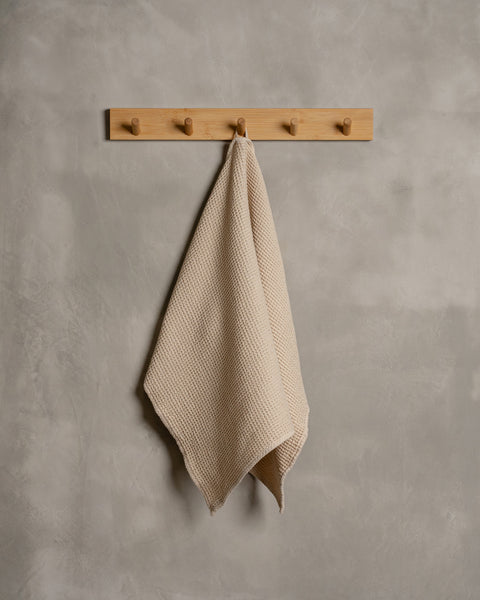 A gray wall with a wooden hanger. In the middle of it hangs a waffle hand washcloth in sand brown color.