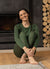 A woman wearing dark green merino wool base layers, sitting down on the floor barefoot by a fireplace.