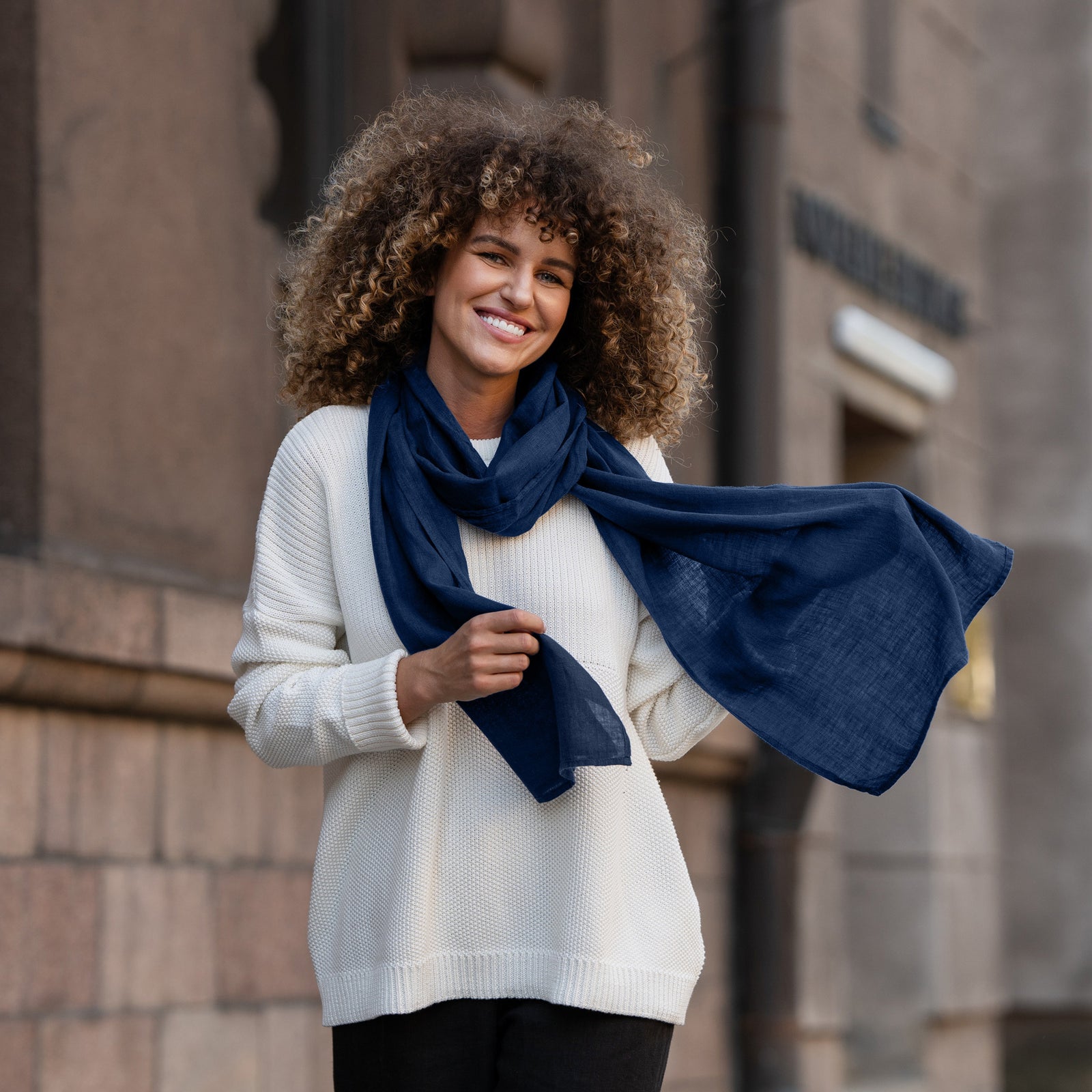 A person standing outdoors wearing a storm blue menique linen scarf draped over the shoulders, with a beige linen dress. The image captures a side profile of the person against a backdrop featuring a gray wall and greenery.