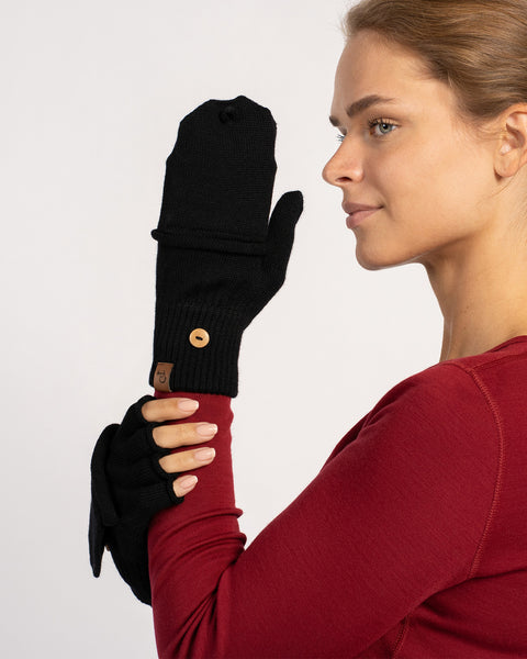 Side-profile of a woman in a red long-sleeve holding up a black convertible mitten (fingerless glove with flap) to show the wooden button detail.