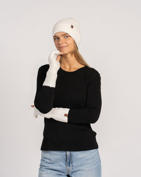 Full-length shot of woman in black top and jeans wearing white beanie and long white wrist warmers, facing camera.