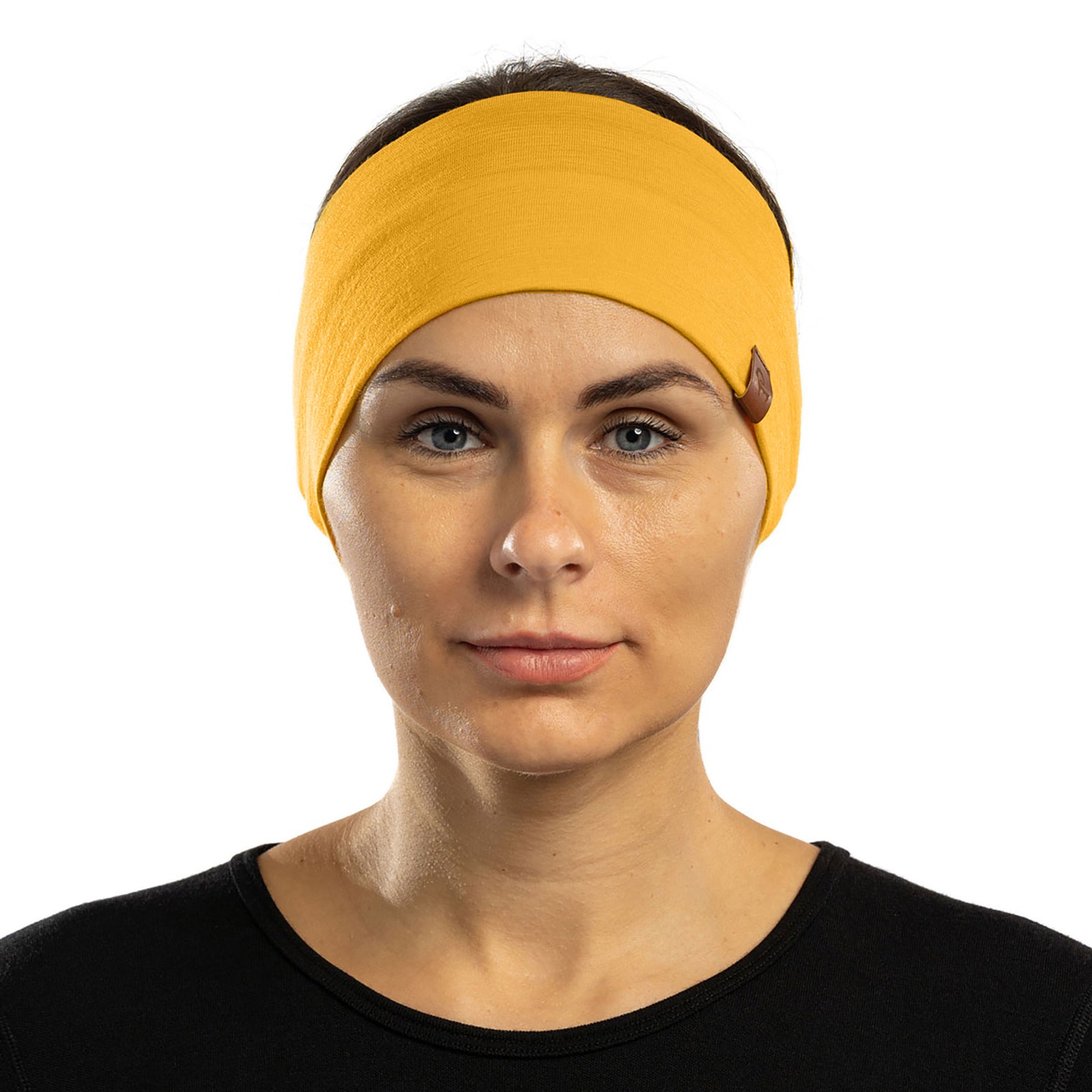 A man and a woman are closely positioned outdoors, both wearing spicy yellow menique merino wool headbands and black long-sleeved shirts. The man, on the left, has short brown hair and a beard and is looking upwards and to his left. The woman, on the right, has light brown hair and is looking upwards with a smile, her hand gently touching the man's face. The background is softly blurred with green and brown tones, suggesting a natural, possibly wooded, environment.