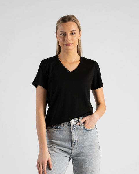 A studio portrait of a blonde woman standing and looking directly at the camera with a subtle smile. She is wearing a black short-sleeved V-neck t-shirt and light wash denim jeans, with one hand casually resting in her front pocket against a plain light grey background.
