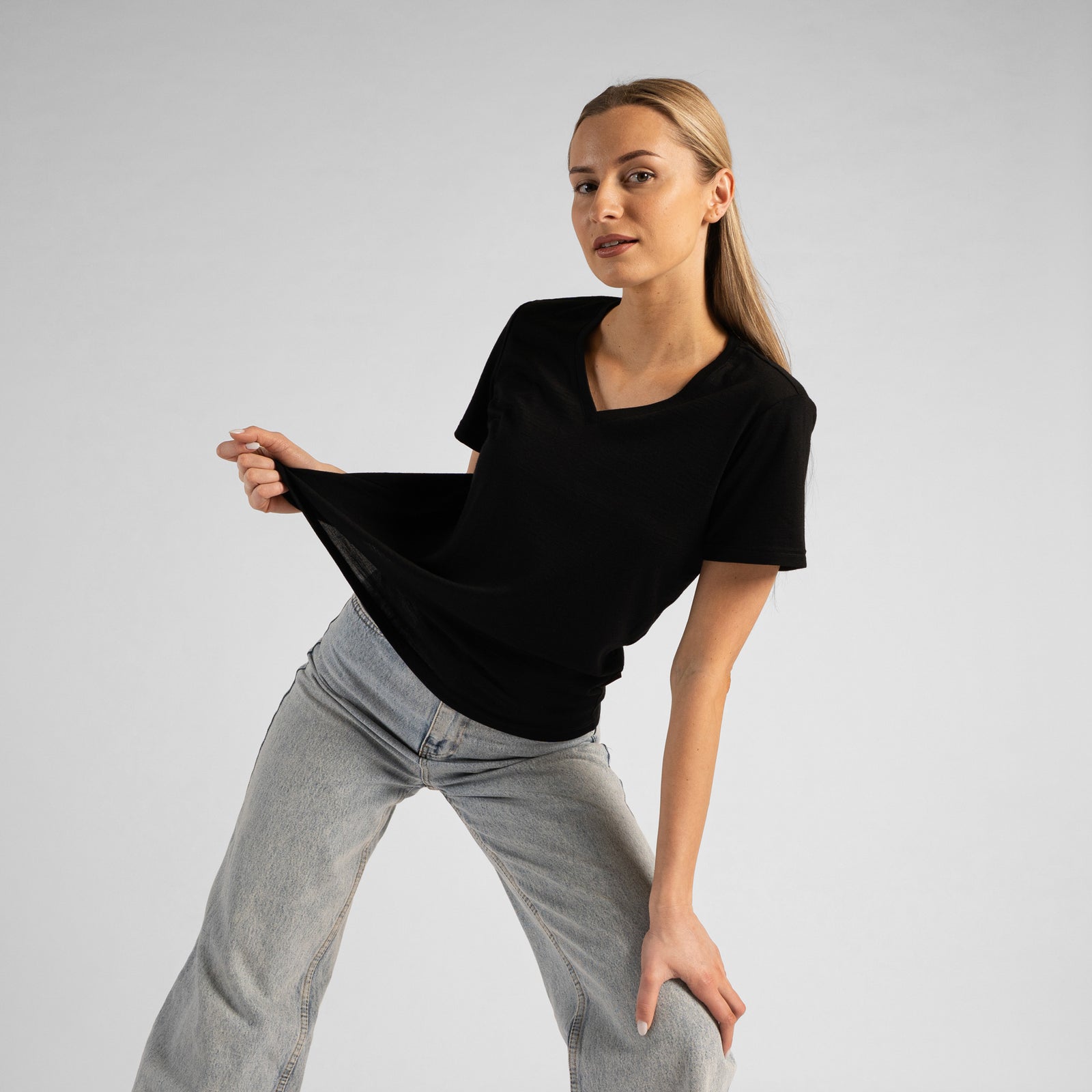A studio portrait of a blonde woman sitting on a stool and looking directly at the camera. She is leaning back slightly, with her hands resting on her lap. She is wearing a regular fit, dusty blue short-sleeved v-neck t-shirt and light-wash blue jeans against a plain light grey background.