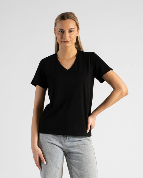 A studio portrait of a blonde woman standing and looking directly at the camera with a subtle smile. She is wearing a black short-sleeved V-neck t-shirt and light wash denim jeans, with one hand resting on her hip against a plain light grey background.