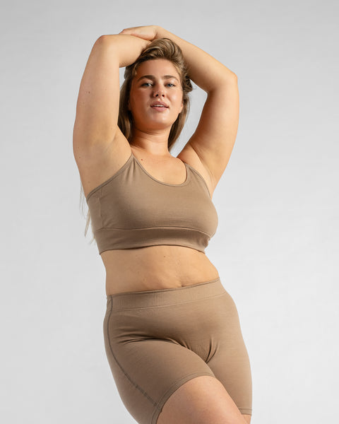 Front three-quarter view of a model with her hands resting on her head, wearing a beige sports bra and matching beige biker shorts.