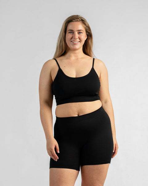 Front view of a smiling model standing with her arms relaxed, wearing a black sports bra with thin straps and black biker shorts.