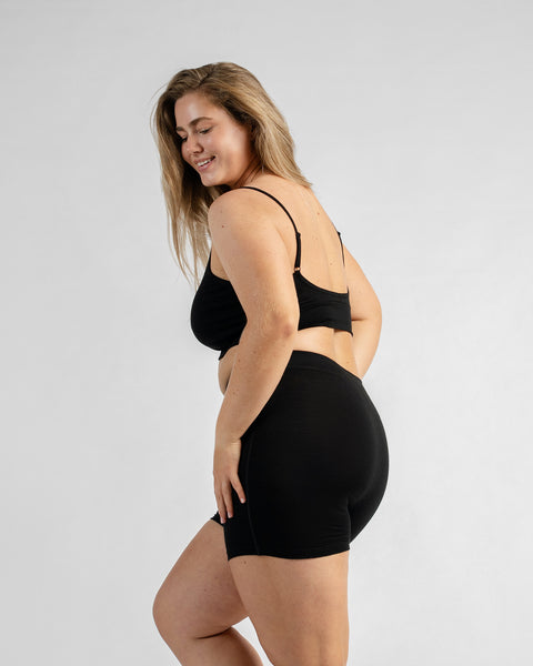 Side-back view of a model looking over her shoulder towards the camera, wearing a black sports bra with thin straps and black biker shorts.