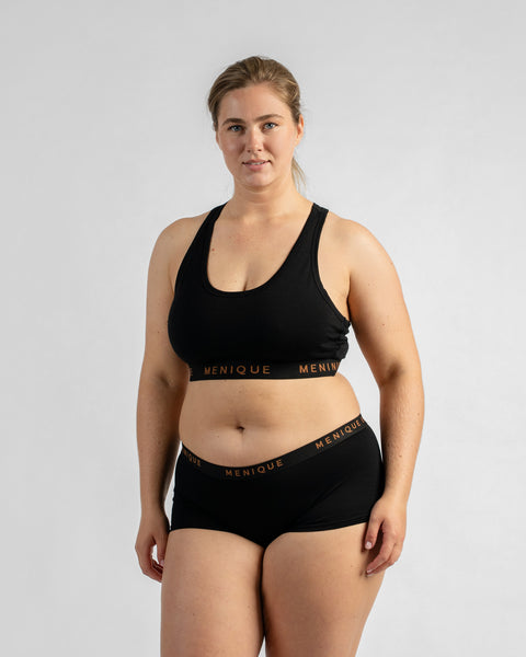 Front view of a model standing with her hands resting lightly on her upper thighs, wearing a black sports bra and matching boy-short underwear, both featuring a black elastic band with text.
