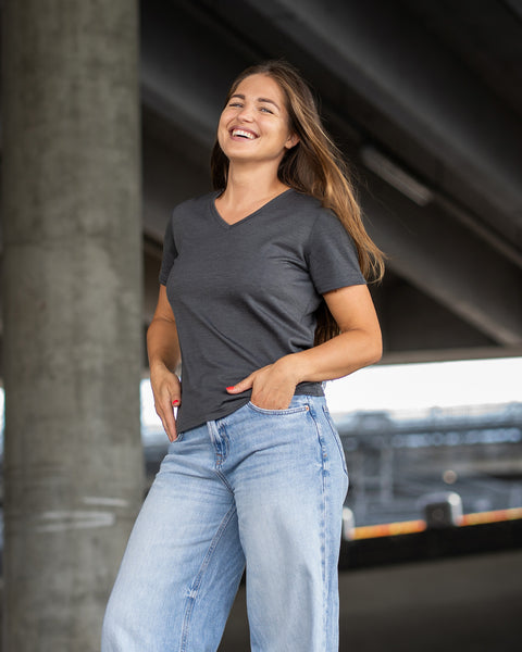 A woman with long brown hair stands outdoors, laughing brightly with her head tilted back. She has her hands tucked into the front pockets of her loose light blue jeans and is wearing a dark grey short-sleeve v-neck t-shirt. The background is a shaded outdoor area beneath a large concrete overpass.