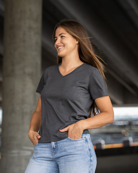 A smiling woman with long brown hair stands outdoors with her hands resting in the front pockets of her light blue jeans, looking off to the left. She is wearing a dark grey short-sleeve v-neck t-shirt. The setting is in the shade underneath a concrete bridge structure.