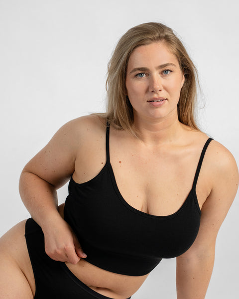 A woman sitting down and looking at the camera, wearing a plain black scoop-neck bralette and black bottoms.
