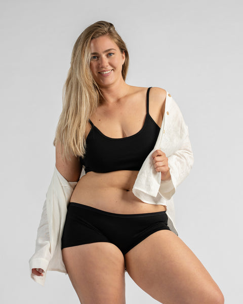 A smiling woman standing while wearing a plain black bralette, black boxer briefs, and an unbuttoned white long-sleeve shirt.