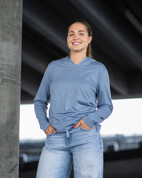 A woman standing in front of a concrete pillar under an overpass, smiling directly at the camera. She is wearing a dusty blue lightweight hoodie and light blue jeans with her hands in her pockets.