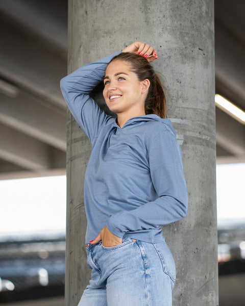 A woman leaning casually against a concrete pillar under a bridge, smiling and looking away to her left. She is wearing a dusty blue hoodie and light blue jeans, with one hand resting against the pillar above her head.