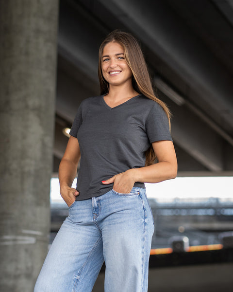 A woman standing outdoors under a concrete overpass, smiling at the camera. She is wearing a charcoal grey short-sleeve v-neck t-shirt and light blue jeans, with her thumbs resting in her front pockets.