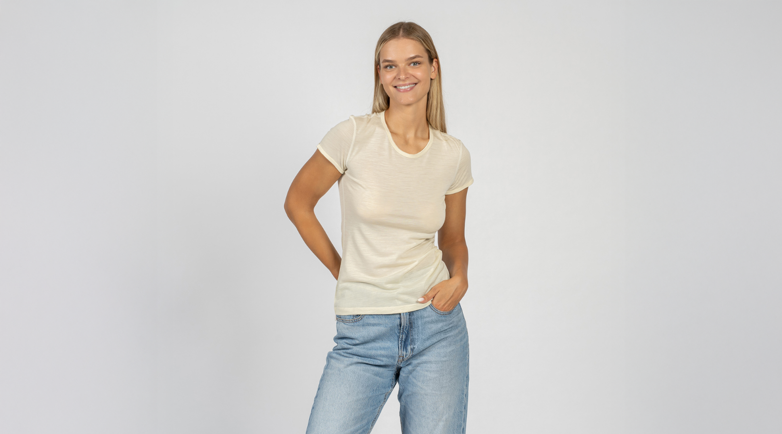A smiling blonde woman standing against a plain light grey studio background, wearing a fitted cream-colored short-sleeved t-shirt and light blue jeans.