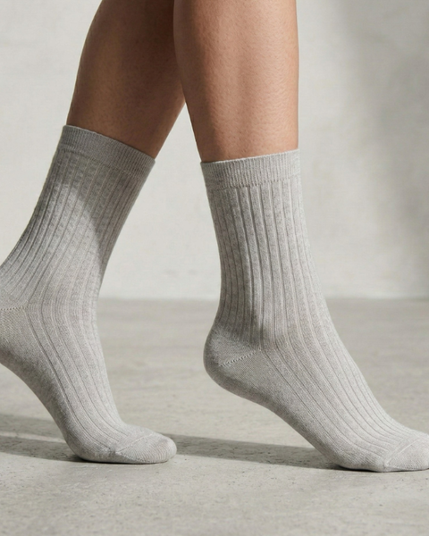 A close-up side view of a person's lower legs and feet wearing light gray ribbed socks. The person is standing on a light gray concrete floor, with one heel slightly lifted. The background is a plain, softly textured light gray wall.