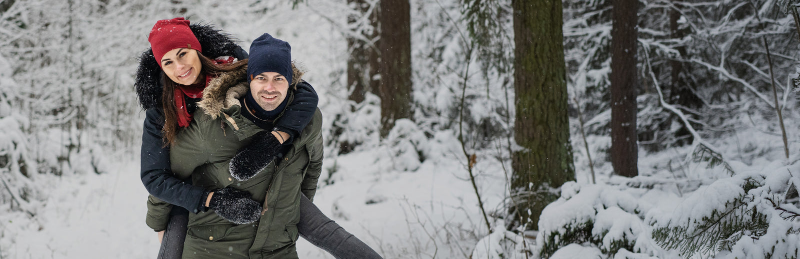 Couple enjoying winter outdoors, woman in red beanie hugging man from behind in snowy forest