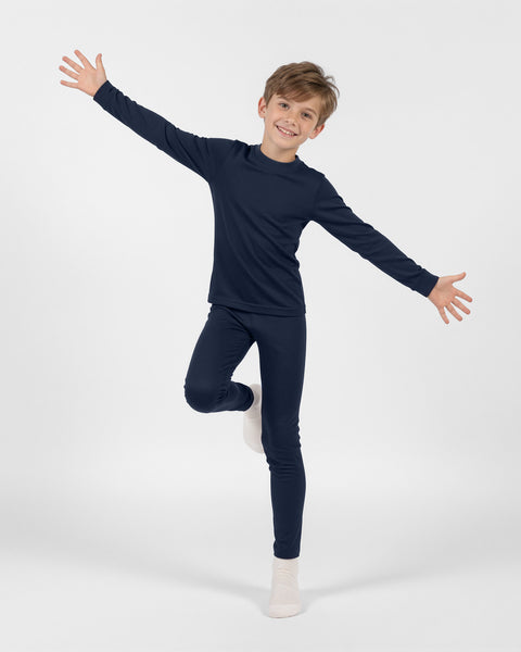 Young boy smiling while balancing on one leg with arms outstretched, wearing a dark navy blue long-sleeve thermal base layer set and white socks against a plain white background.