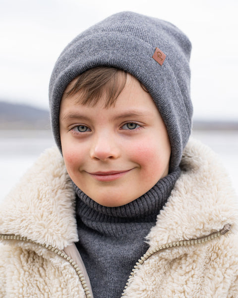 A young child with blue eyes and light brown hair smiles softly at the viewer while wearing a grey knit beanie with a brown label, a grey turtleneck, and a cream-colored sherpa jacket. The background is a blurred, wintry landscape with what appears to be water.