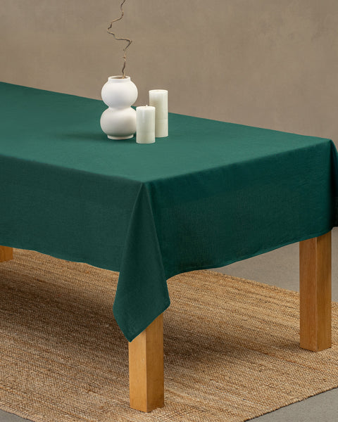 The menique linen tablecloth in dark green color, draping over a wooden table, with a white modern vase on top of it, and two different height white candles by the sides.