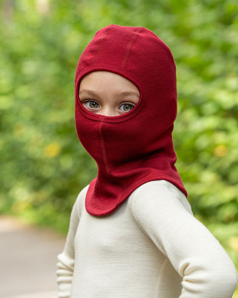A young child wearing a royal cherry knit balaclava and a cream-colored shirt looks over their shoulder at the camera, with a background of blurred green foliage.