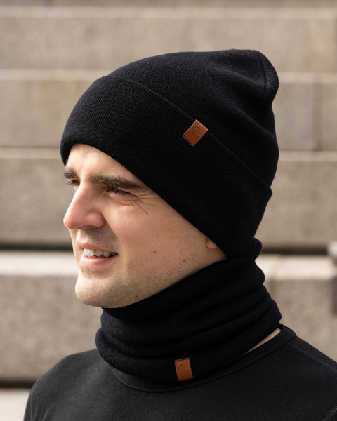 A person with light skin and dark hair is shown in profile, smiling slightly. They are wearing a black knit beanie with a small brown tag and a matching black knit neck gaiter, also with a small brown tag. They are wearing a black long-sleeved shirt. The background appears to be concrete steps or a similar textured surface in an outdoor setting.
