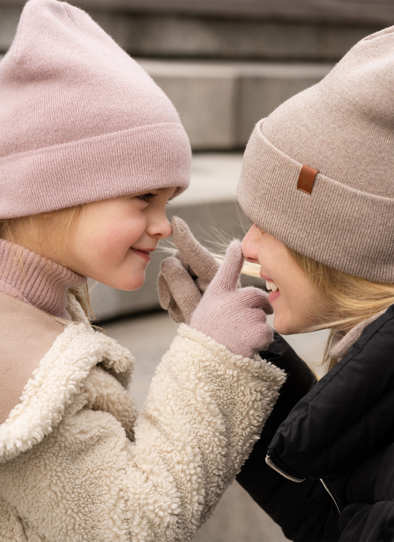 A woman with her daughter playfully touching each other's nose with the fingertip of their merino wool gloves. the mother is wearing a dickie, beanie hat and gloves in creamy beige, while the daughter is wearing the same accessories in kids' sizing and dusty pink color.