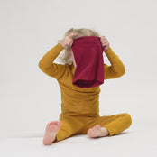 A happy child with light hair, wearing a yellow colored long-sleeved top and matching pants, and a royal cherry neck warmer, sits cross-legged on a white surface. The child is smiling broadly at the camera.