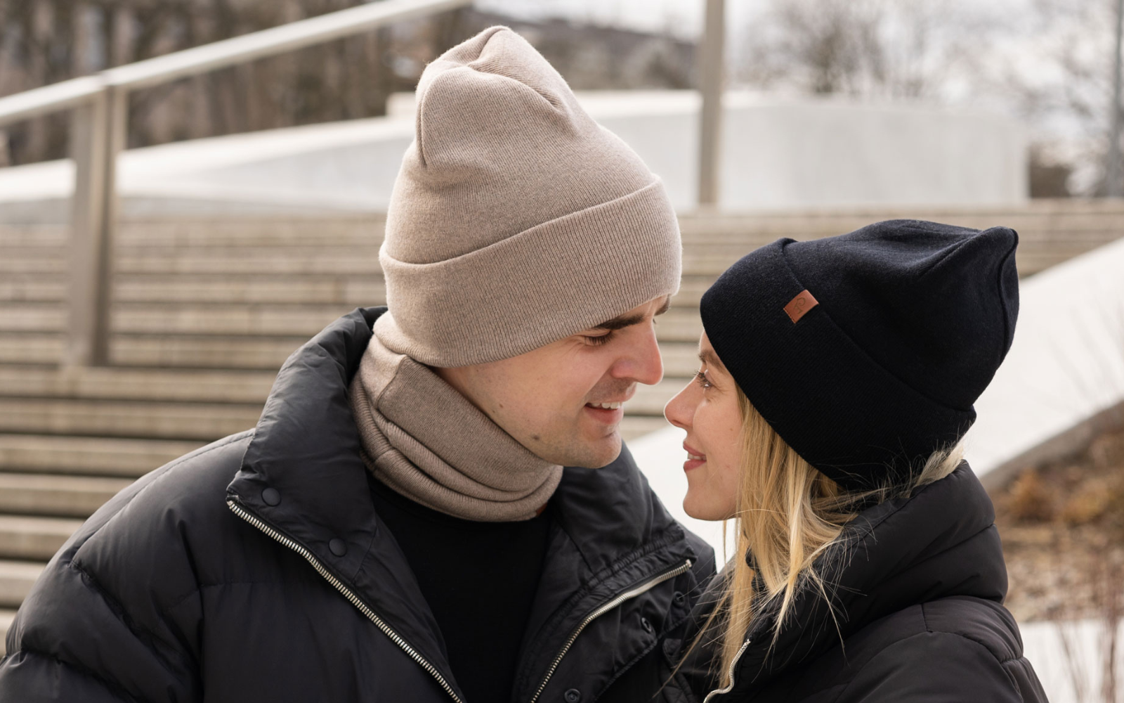 A man and a woman near each other, wearing winter accessories. Both of them are wearing a beanie hat and dickie neck warmer: the woman in black, and the man in creamy beige color.