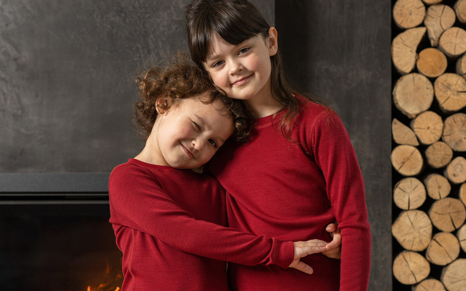 Two girls wearing royal cherry color 250gsm merino wool clothing sets, standing by a fireplace.