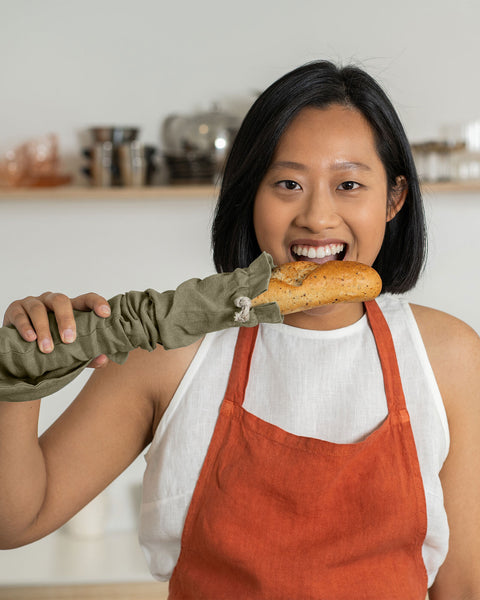A woman trying to bite a baguette that is stored in a linen baguette bag in stone green color.