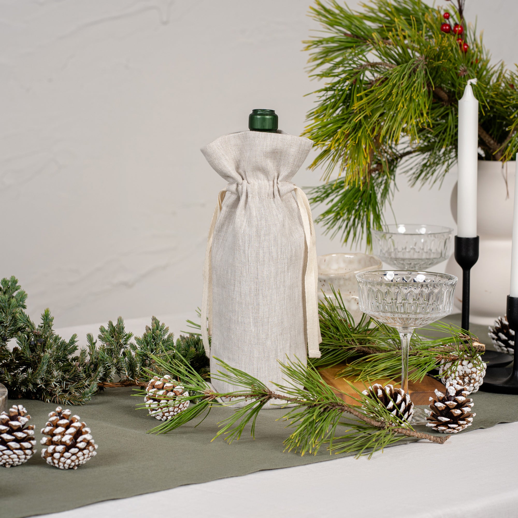 The menique linen wine bottle gift bag in pure red color. The bottle is put on top of a Christmas table, decorated with pine cones, fir branches, and other natural elements. The table is covered with a pure white tablecloth and a stone green table runner. There are candles and glasses on the table as well. 