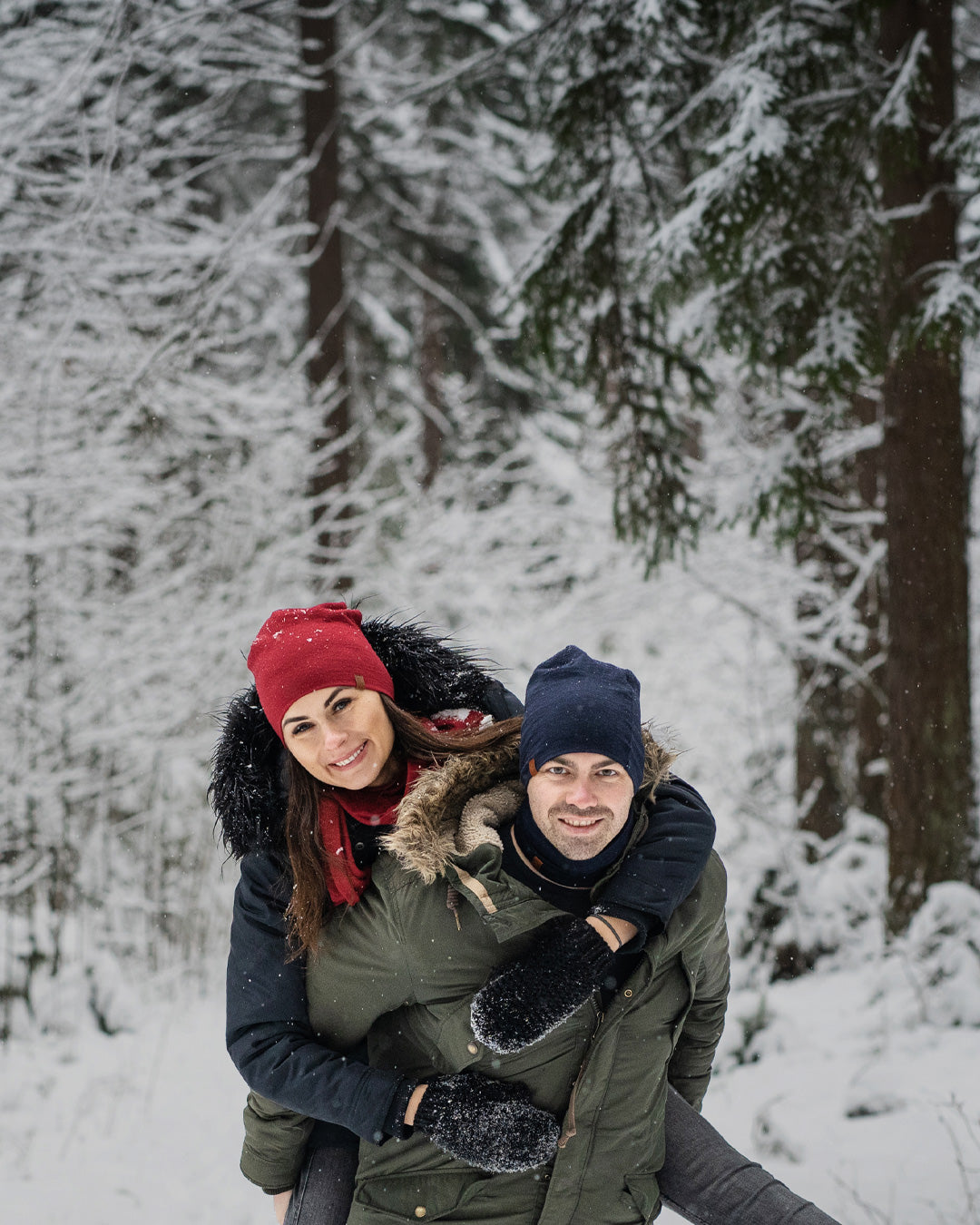 Couple enjoying winter outdoors, woman in red beanie hugging man from behind in snowy forest
